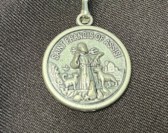 May include: A silver-colored pendant with a loop at the top. The pendant features an image of Saint Francis of Assisi with animals and the text "SAINT FRANCIS OF ASSISI" around the edge. The pendant is round.