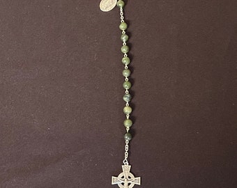 Puede incluir: Un rosario con cuentas verdes y un colgante de cruz celta plateada. El rosario también presenta una pequeña medalla plateada y un llavero. Las cuentas son una mezcla de verde y gris.