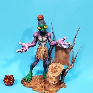 May include: A fantasy figurine of a purple alien-like creature with large yellow eyes, green feet, and a small orange hair bun. It stands next to a brown rock with a handprint and a small banner. A red and gold die is in the corner.