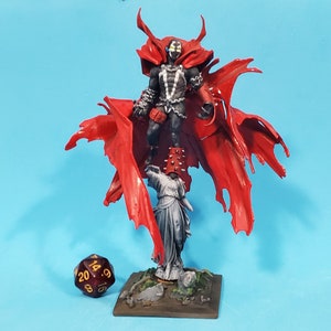 Puede incluir: Una figura miniatura pintada de rojo y negro de un superhéroe con una capa roja y una calavera blanca y negra en el pecho. La figura está de pie sobre una base con una estatua gris y un dado de 20 caras marrón.