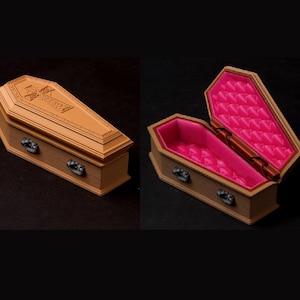 May include: A miniature coffin, shown closed and open, with a detailed cross design on the lid. The coffin is brown with dark metal handles and a bright pink interior. The open coffin reveals a quilted pink lining.