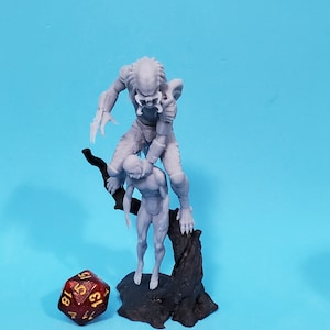May include: A gray plastic miniature figurine of a Predator holding a human figure, both standing on a gray plastic tree base. A red 20-sided die with gold numbers is in the foreground.