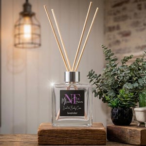 May include: A square clear glass reed diffuser bottle with a silver cap and three wooden reeds. The black label features the text "NF Milena Flowers Soul & Body Care" and "lavender" in white and pink lettering, centered on the front of the bottle.