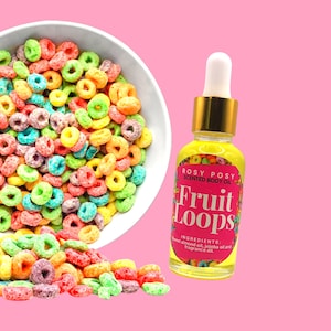 Puede incluir: Una pequeña botella de vidrio de aceite corporal perfumado con tapa dorada y una etiqueta colorida que dice "ROSY POSY SCENTED BODY OIL Fruit Loops INGREDIENTS: Sweet almond oil, jojoba oil and fragrance oil". La botella está sobre un fondo rosa con un tazón de cereal colorido que se derrama.