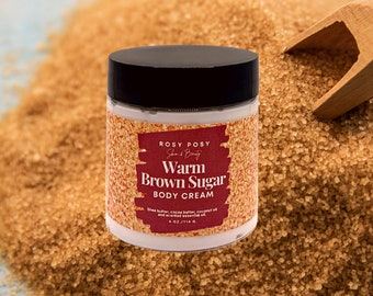 May include: A white jar of body cream with a black lid. The jar is labeled "ROSY POSY Skin & Beauty Warm Brown Sugar Body Cream" and has the text "Shea butter, cocoa butter, coconut oil and scented essential oil 4 oz/114 g". The jar is sitting on a pile of brown sugar.