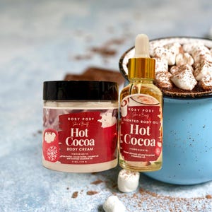 May include: A jar of Hot Cocoa body cream and a bottle of Hot Cocoa scented body oil. The products are labeled with the brand name "Rosy Posy She & Beauty" and are red and white. A blue mug of hot cocoa with marshmallows is in the background.