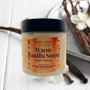 Puede incluir: Un tarro blanco con tapa negra y una etiqueta amarilla que dice "ROSY POSY Skin & Beauty Warm Vanilla Sugar BODY CREAM INGREDIENTS: Shea butter, cocoa butter, coconut oil and scented essential oil 4 OZ./114 G."