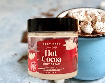 May include: A clear jar of Rosy Posy Hot Cocoa Body Cream with a black lid. The label is red with white text and a graphic of a mug with marshmallows. The jar is 4 oz. and is next to a blue mug of hot cocoa with marshmallows.