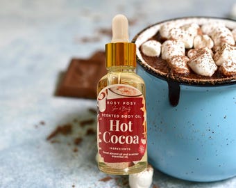 May include: A clear glass bottle of "Hot Cocoa" scented body oil with a gold dropper. The label is red and white. A blue mug filled with hot chocolate and marshmallows is in the background. Chocolate pieces and marshmallows are scattered around.