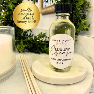 以下が含まれることがあります： 黒い蓋が付いた透明なガラス瓶に、黄色の液体が入っています。瓶には白いラベルが付いており、"ROSY POSY Skin & Beauty LUXURY Escape HOME DIFFUSER OIL 2 OZ."と書いてあります。
