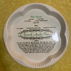 May include: White ceramic plate with a scalloped edge, featuring a recipe for French Apple Flan. The text includes ingredients like flour, butter, and apples, along with instructions. The plate has a decorative floral design.