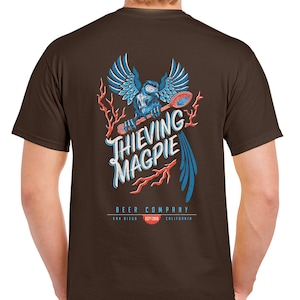 Op de afbeelding: Bruin T-shirt met een blauwe en rode illustratie van een ekster die een lepel vasthoudt. De tekst "Thieving Magpie Beer Company" is onder de illustratie gedrukt. De tekst "San Diego Est. 2018 California" is onder de bedrijfsnaam gedrukt.