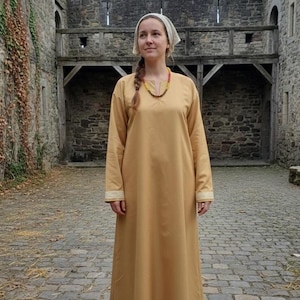 May include: A long, mustard-colored medieval dress with long sleeves and a simple neckline. The dress has white and gold trim on the cuffs. A woman is wearing the dress, a white head covering, and a colorful beaded necklace. The background is a stone castle.