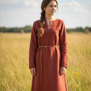 May include: A long-sleeved, rust-colored dress with a V-neckline and a brown belt. The dress is made of a soft fabric and has a flowing skirt. The woman is standing in a field of tall grass.