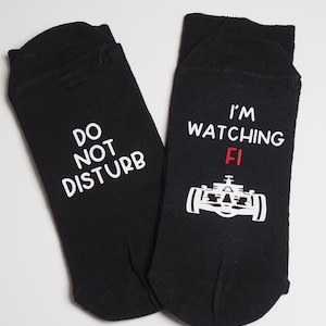 May include: A pair of black socks with white text. One sock says "Do Not Disturb" and the other says "I'm Watching F1" with a white image of a racing car and the letter "F" in red.