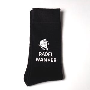 May include: A black crew sock featuring a white graphic of a padel racket with a ball above it. Below the graphic, the text PADEL WANKER is printed in a bold, white, sans-serif font. The sock has a ribbed cuff at the top and a smooth knit texture.