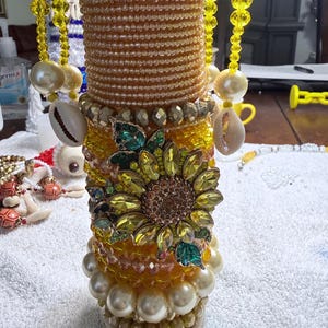 May include: A decorative, cylindrical object adorned with layers of yellow and gold beads, pearls, and seashells. A large, jeweled sunflower is centered on the object. Strands of yellow beads and large pearls hang from the top.