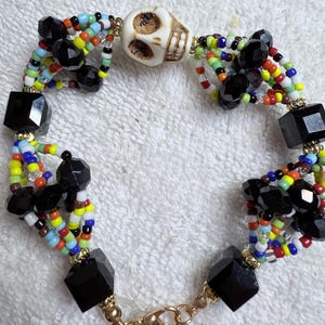 May include: A beaded bracelet featuring a central skull bead and black faceted beads. The bracelet incorporates colorful seed beads in various hues, with gold-toned metal accents and a clasp. The design is intricate and detailed.
