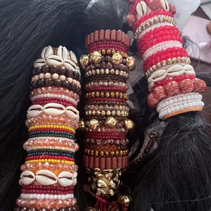 May include: Three decorative hair wraps, each featuring a unique design. One wrap has a combination of brown, white, and orange beads with seashells. Another has gold beads and a burgundy color. The third wrap has red and white beads with seashells.