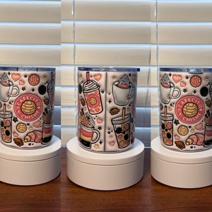 May include: Three white stainless steel travel mugs with pink and brown designs. The mugs feature a variety of images including hearts, pastries, coffee cups, and the text "Cafecito y Chisme".