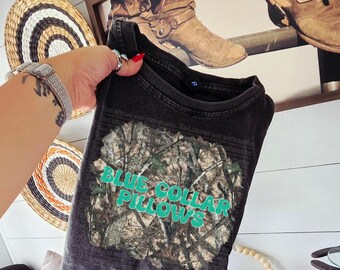 May include: Dark gray t-shirt with a camouflage design and the words "BLUE COLLAR PILLOWS" in turquoise. The shirt is held up in front of a rustic background with a framed picture of cowboy boots.