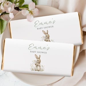 May include: Two white chocolate bar wrappers with gold edges, featuring a watercolour illustration of a bunny holding flowers. The text "Emma's Baby Shower" is written in a delicate script. The bars are on a white plate with floral accents.