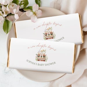 Puede incluir: Dos envoltorios de barras de chocolate blancos con bordes dorados, con el texto "a new chapter begins" y "Sophia's Baby Shower". Una ilustración floral de libros y flores está centrada en el envoltorio. Las barras descansan sobre un plato blanco con flores rosas.