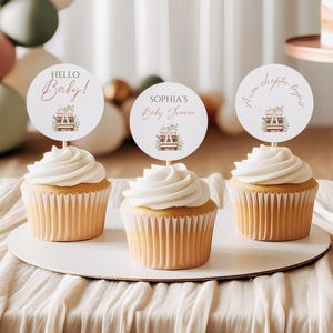 Peut inclure: Trois cupcakes avec glaçage blanc et décorations. Les décorations rondes affichent les textes "Hello Baby!", "Sophia's Baby Shower" et "A new chapter begins". Chaque décoration présente un motif floral et de livre.