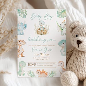 Puede incluir: Una invitación para un baby shower con temática de dinosaurios. La invitación presenta ilustraciones en acuarela de varios dinosaurios en colores pastel, junto con el texto "Baby Boy" y "hatching soon". La invitación incluye detalles del evento.