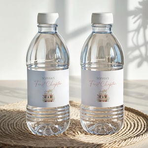 May include: Two clear plastic water bottles with white caps. Each bottle has a label that reads "Sophia's First Chapter" in a script font, with a floral and book illustration. The bottles are on a woven mat.