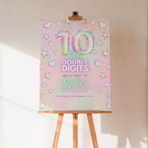 Könnte beinhalten: Ein Willkommensschild zum Geburtstag auf einer Staffelei. Das Schild hat einen pastellfarbenen Regenbogenhintergrund mit Herzen und Sternen. Die Zahl 10 steht in der Mitte, darunter die Worte "hello double digits" und "Welcome to Sophie's 10th Birthday". Das Datum ist Samstag, der 27. September.