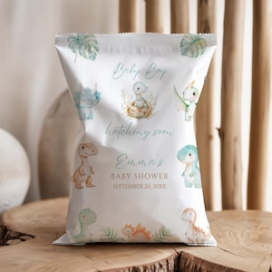 Op de afbeelding: Witte baby shower traktatiezak met aquarel illustraties van dinosaurussen en tropische bladeren. De tekst "Baby Boy", "hatching soon", "Emma's Baby Shower" en de datum staan op de zak.