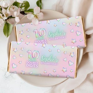 Puede incluir: Dos barras de chocolate con bordes dorados, envueltas en un diseño holográfico pastel. Los envoltorios presentan el texto "10 hello DOUBLE DIGITS Sophie's 10th BIRTHDAY" con gráficos de estrellas y corazones. Las barras están sobre un plato blanco.