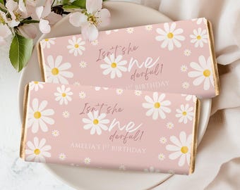 Op de afbeelding: Twee roze chocoladereepverpakkingen met een madeliefjespatroon en goudkleurige randen. De tekst "Isn't she wonderful!" en "Amelia's 1st Birthday" zijn gedrukt. De repen liggen op een wit bord met bloemen.