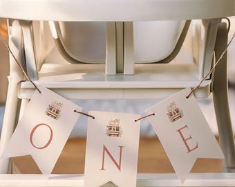 May include: A white banner spelling "ONE" in a soft red font, strung across a high chair. Each banner piece has a floral illustration. The high chair is white and has a neutral colour scheme.