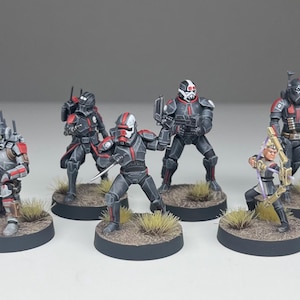 May include: A set of six miniature figurines featuring armored characters in black and red tactical gear. The figures are posed in various combat stances on circular bases with textured ground and small tufts of grass, including one archer.