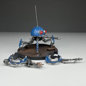 May include: A blue and silver miniature robot with a dome-shaped head, two red circular eyes, and a thin antenna. The robot has three articulated legs and a forward-facing weapon. Two additional detached blue and silver arm components lie in front.