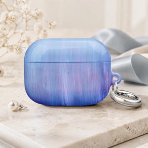 May include: A protective case for wireless earbuds featuring a smooth, marbled finish with blended shades of light blue, violet, and white. The case includes a small side loop with a polished silver-toned metal ring attached for easy carrying.