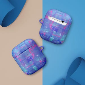 Könnte beinhalten: Zwei AirPod-Hüllen mit abstraktem Design. Die Hüllen weisen eine lebendige Mischung aus Blau, Lila und Pink auf, mit einem strukturierten, bemalten Aussehen. Jede Hülle hat eine kleine Schlaufe und einen Klappdeckel. Der Hintergrund ist in Beige und Blau geteilt.