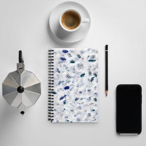 May include: A white notebook with a blue and gray abstract pattern, a black pencil, a silver coffee pot, a white coffee cup with a saucer, and a black smartphone.