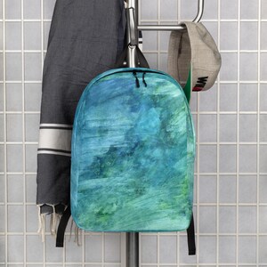 May include: A backpack with a watercolor design in shades of blue and green hangs on a metal rack. The backpack has black zippers and straps. A gray towel with white stripes and a baseball cap are also hanging on the rack.