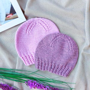 May include: Two knitted baby hats in shades of pink and purple. The hats are made of soft yarn and feature a ribbed brim and a decorative stitch pattern on the crown. The hats are displayed on a neutral-colored fabric.