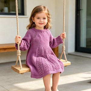 Può includere: Una bambina con un vestito viola lavorato a maglia a maniche lunghe con un design dettagliato sulla gonna, seduta su un'altalena di legno. L'abito ha uno scollo tondo e polsini delle maniche intricati. L'altalena è sospesa da una corda.