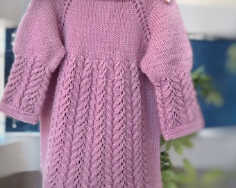 May include: A pink knitted baby dress with a delicate lace pattern. The dress has long sleeves and a round neckline.