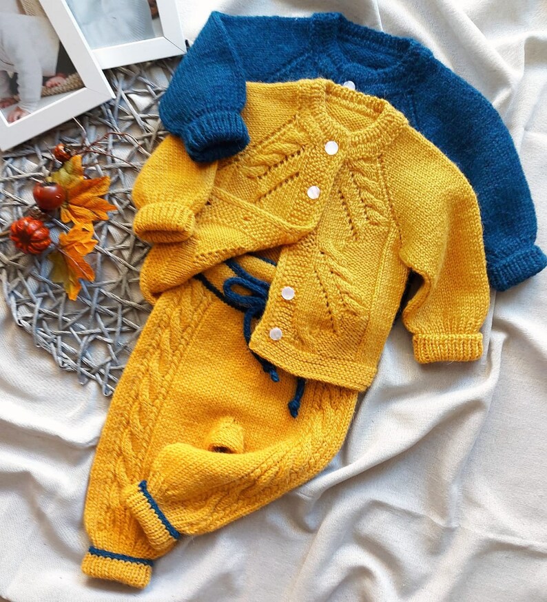 K&ouml;nnte beinhalten: Ein gelber gestrickter Baby-Pullover und eine Hose mit einem blauen gestrickten Cardigan dar&uuml;ber. Der Pullover und die Hose haben ein Zopfmuster und der Cardigan ein Blatt-Muster.