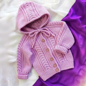 May include: A light purple, hand-knitted baby cardigan and matching hooded hat set. The cardigan features a cable knit design, wooden buttons, and a tie closure. The set is displayed on a white and purple background.