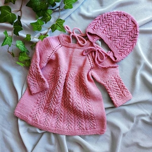 Könnte beinhalten: Ein rosa gestricktes Babykleid und ein passendes Mützchen-Set. Das Kleid hat ein detailliertes Blattmuster auf dem Oberteil und den Ärmeln, mit einem runden Ausschnitt und langen Ärmeln. Die Mütze hat ein ähnliches Blattmuster.