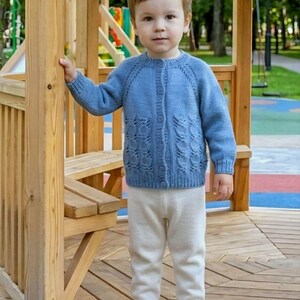 Könnte beinhalten: Eine blaue, langärmelige Strickjacke mit durchgehender Knopfleiste und einem strukturierten Blattmuster an den unteren Vorderteilen, kombiniert mit einer schlichten, cremeweißen Strickhose und weißen Sneakern mit Klettverschluss.