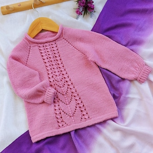 May include: A light pink knitted sweater with a central openwork design. The sweater has long sleeves and a ribbed hem and cuffs. It is displayed on a wooden hanger against a backdrop of white and purple fabric.