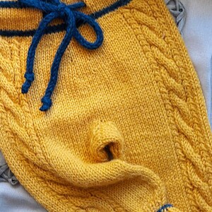 K&ouml;nnte beinhalten: Gelbe gestrickte Babyhose mit blauem Kordelzug und blauem Besatz. Die Hose hat ein Zopfmuster.